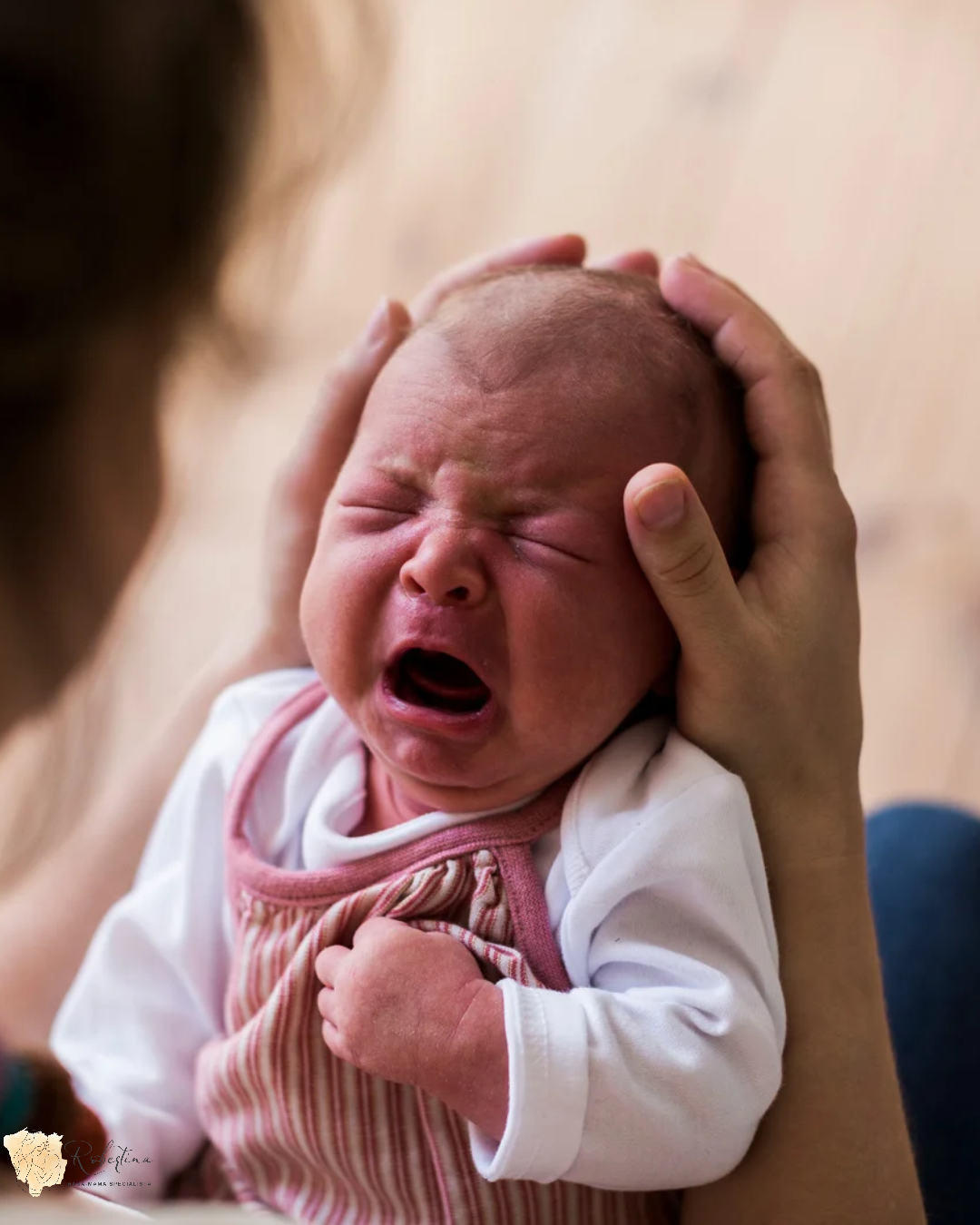 Síró újszülöttet tartanak a karokban, a baba sírva csukott szemmel, kéz a fején.